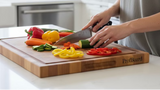A Chef knife with a silver stainless steel blade and a black handle secured by three silver rivets, lying horizontally on a glossy reddish-brown wooden surface.