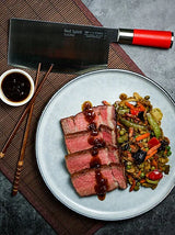 Slicing chef knife with red handle on plate with sliced meat and vegetables.