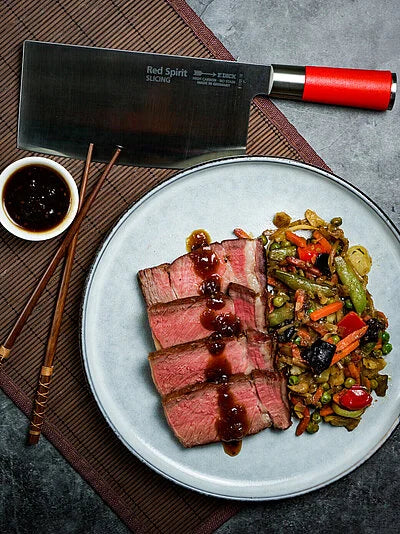 Slicing chef knife with red handle on plate with sliced meat and vegetables.
