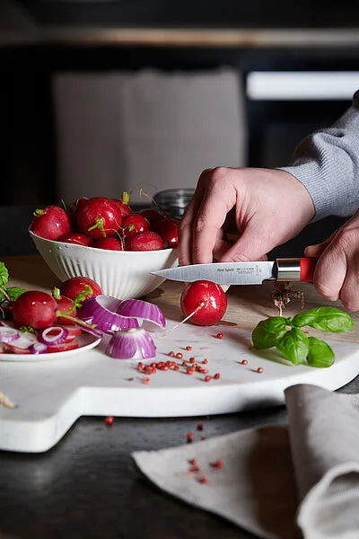 Red-handled paring knife in use slicing radishes on a white cutting board