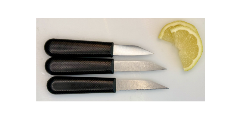 Three small knives with black handles on a white surface next to a lemon wedge.